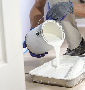 Peinture blanche : Préparer le bac pour une rénovation réussie. Peintre en gants bleus versant de la peinture blanche fraîche d'un pot dans un bac à rouleau pour préparer la rénovation intérieure.
