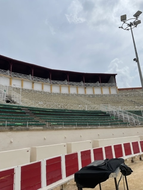 Gradins d'une arène avec sièges verts, barrière rouge et blanche et toit à arcades sous un ciel couvert.