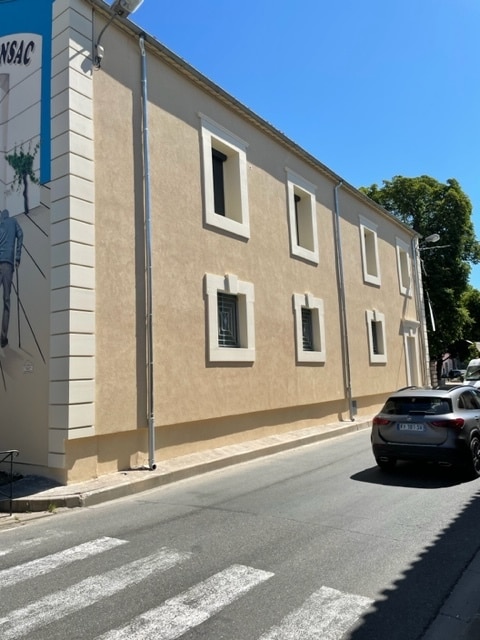 Façade beige d'un bâtiment avec fresque murale 'NSAC', fenêtres blanches et voiture grise sous un ciel bleu.