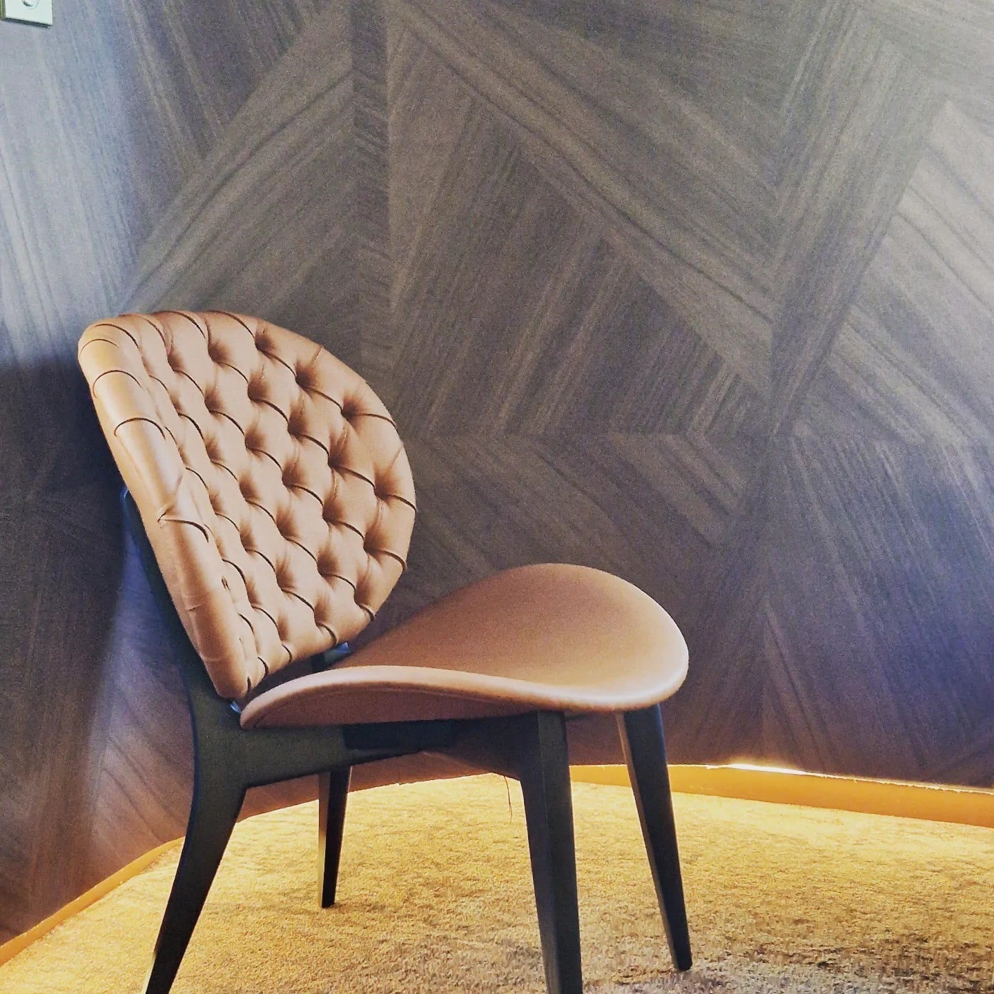 Fauteuil moderne en cuir fauve avec dossier capitonné devant un mur à motifs géométriques gris.