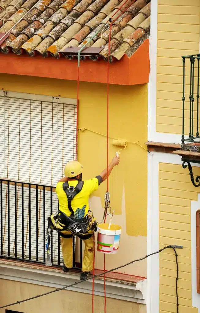 Peinture de façade en hauteur : ouvrier cordiste au travail Un ouvrier en harnais peint un mur extérieur jaune avec un rouleau, suspendu par des cordes depuis un toit en tuiles.