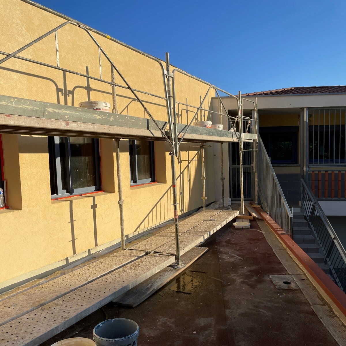 Échafaudage sur terrasse pour rénovation de façade jaune, fenêtres noires et pots de peinture sur chantier.