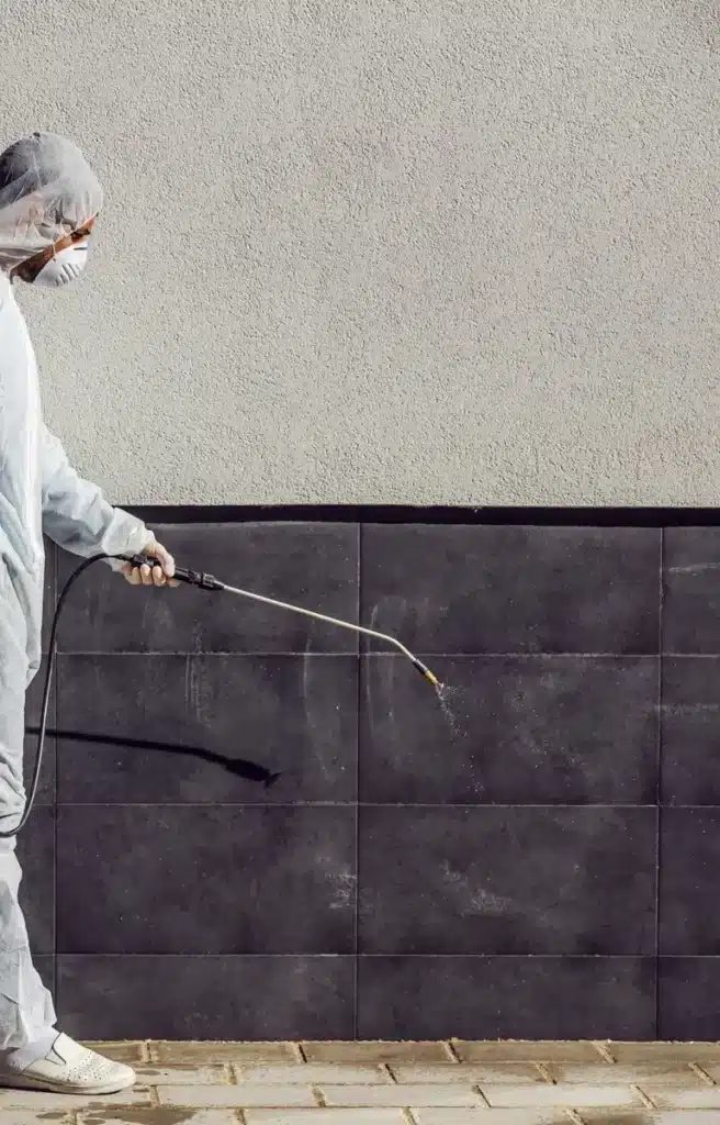 Désinfection Professionnelle et Traitement de Surface Technicien en combinaison de protection pulvérisant un traitement désinfectant sur un mur extérieur carrelé.