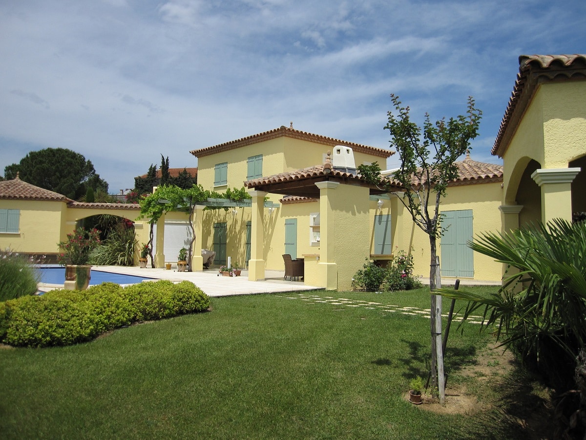 Villa méditerranéenne jaune avec piscine, volets verts et jardin paysager sous un ciel bleu ensoleillé.